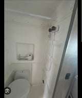 Compact white RV rental wet bath featuring a toilet, shower controls with hose, and a small shelf holding toilet paper.