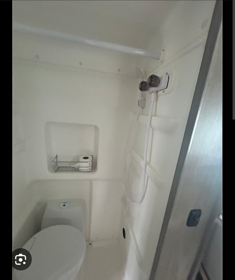 Compact white RV rental wet bath featuring a toilet, shower controls with hose, and a small shelf holding toilet paper.