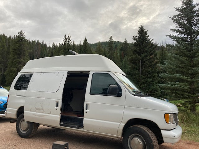 Spacious white high-top camper van, an ideal RV rental for exploring forests. Open door shows cozy interior for van life.