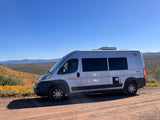 A silver camper van (RV rental) on a dirt road, amidst orange wildflowers with hills under a clear blue sky.