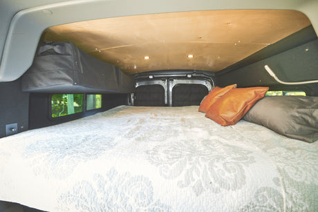 Cozy RV rental camper van interior with a large bed, patterned quilt, leather and fabric pillows, and overhead storage.
