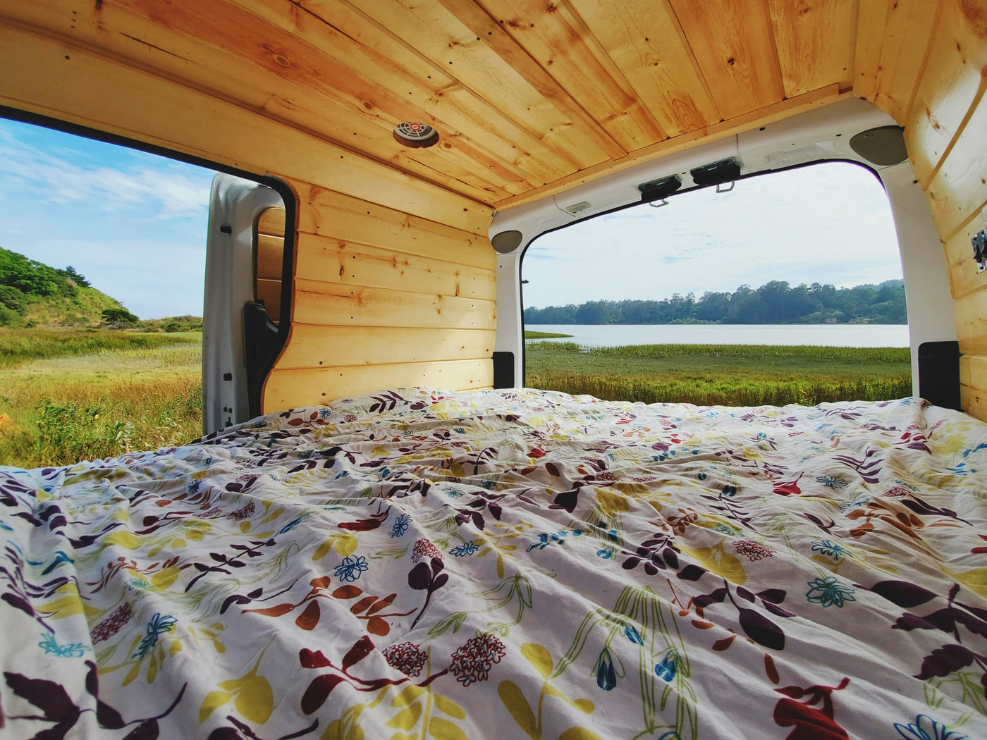 Inside a custom rv rental van, a comfy bed faces open doors with a beautiful view of a lake and grassy hills. — photo 10