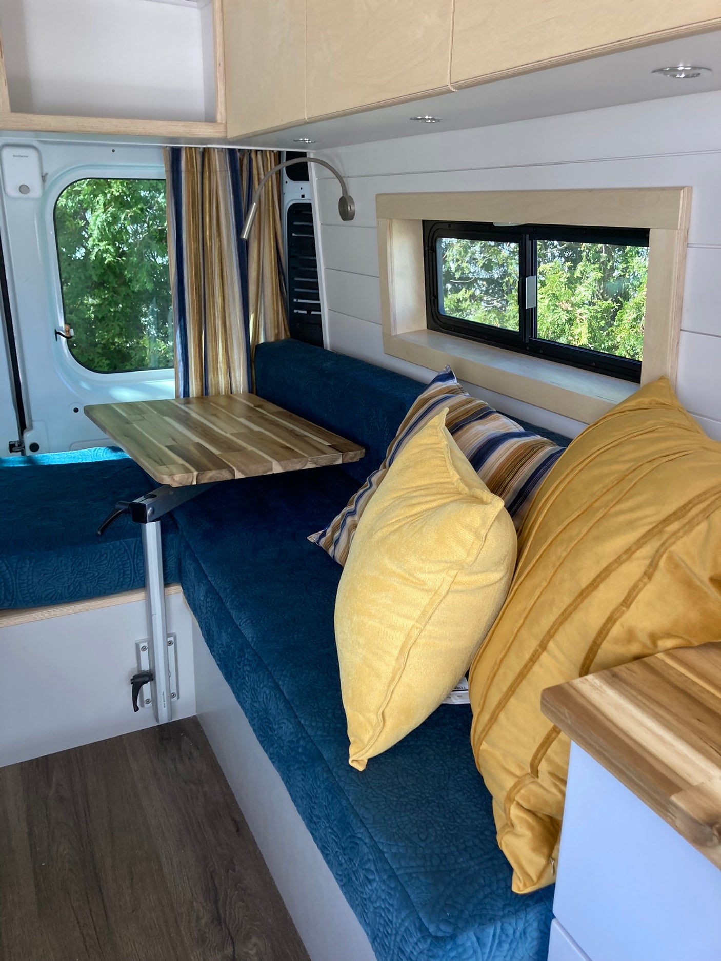 This inviting RV rental camper van interior features blue upholstered seating, a wood table, and sunlit windows with tree views.