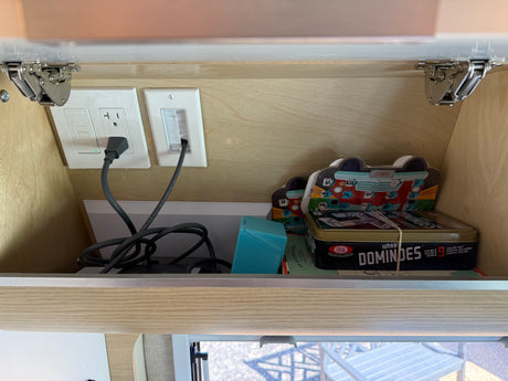 Inside an RV rental overhead cabinet: electrical outlets, cables, and travel games like Dominoes for entertainment on the go.