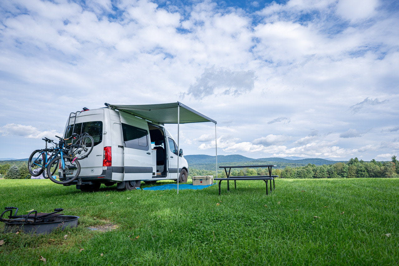 Enjoy an RV rental experience! White camper van with bikes in a grassy field, awning, picnic table & mountain views.