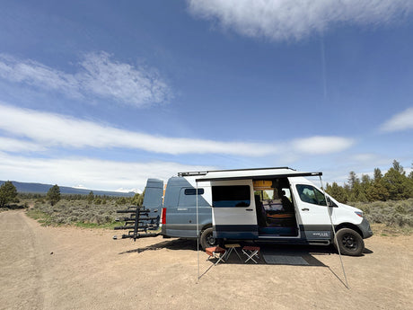 RV rental van with awning & chairs on dirt road, a vast landscape of sagebrush & distant snow-capped mountains.
