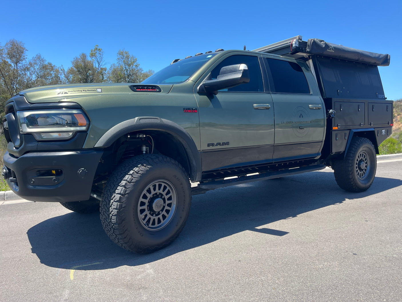 Olive green RAM truck RV rental, modified for overlanding with a custom utility bed, awning, and large off-road tires.