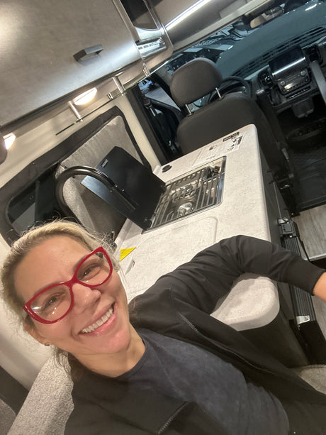 Smiling woman in red glasses inside an RV rental. View from living area shows compact kitchen and front cockpit.