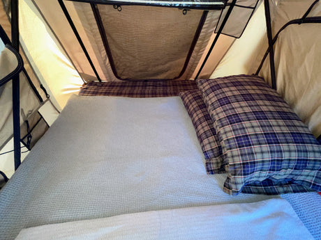 RV rental sleeping space: A cozy bed with a light grey blanket and blue plaid pillows inside a well-lit rooftop tent.