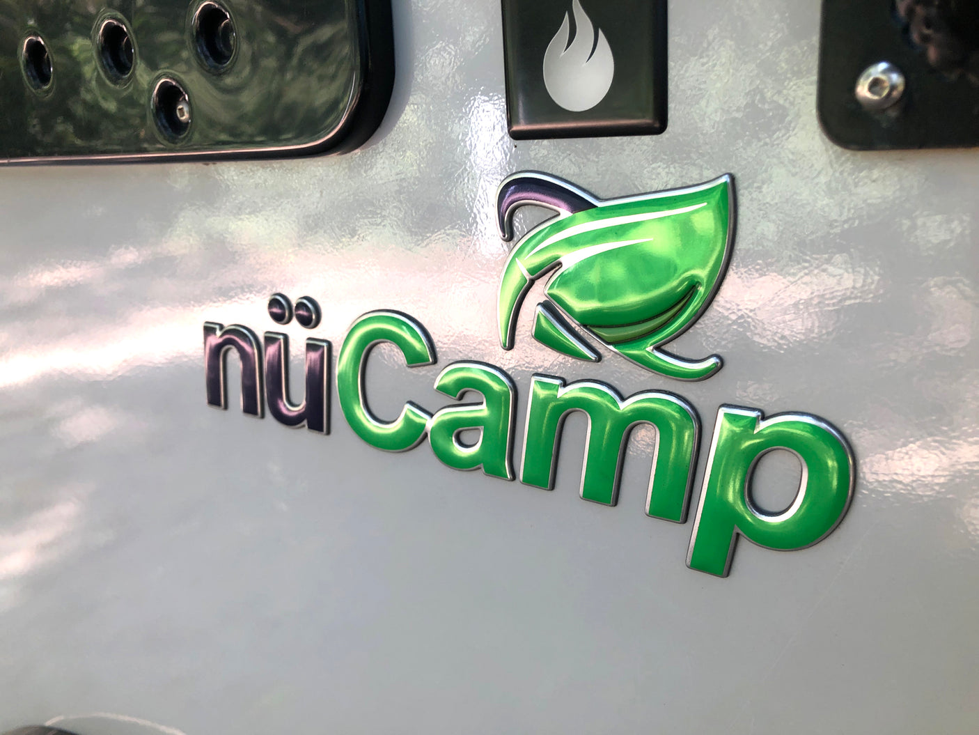 Close-up of a nüCamp RV logo on a white surface, with metallic green "Camp" text and a leaf graphic, ideal for an RV rental.
