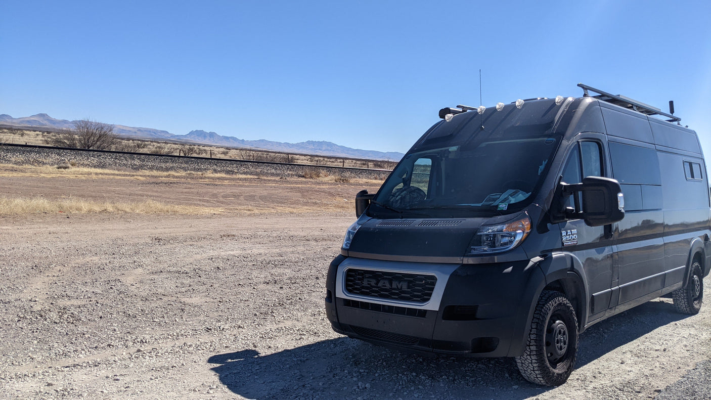 Adventure-ready dark gray Ram ProMaster camper van on a gravel road by railroad tracks & mountains. Perfect RV rental for off-grid travel.