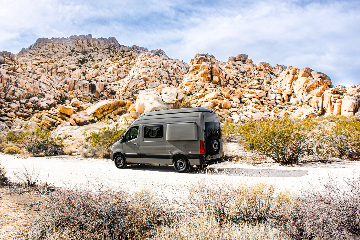 A grey RV rental is parked on a dirt road amidst large desert rock formations under a cloudy sky.