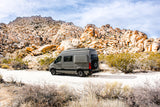 A grey RV rental is parked on a dirt road amidst large desert rock formations under a cloudy sky.