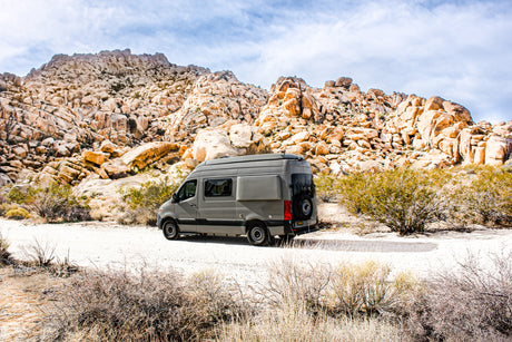A grey RV rental is parked on a dirt road amidst large desert rock formations under a cloudy sky.