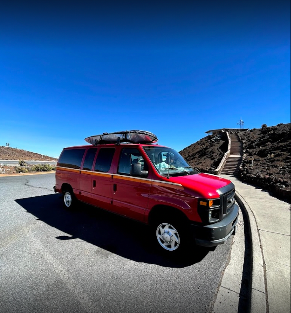 Red **RV rental** van with surfboard on roof, parked on a mountain road with steps leading to a scenic viewpoint under a clear blue sky.