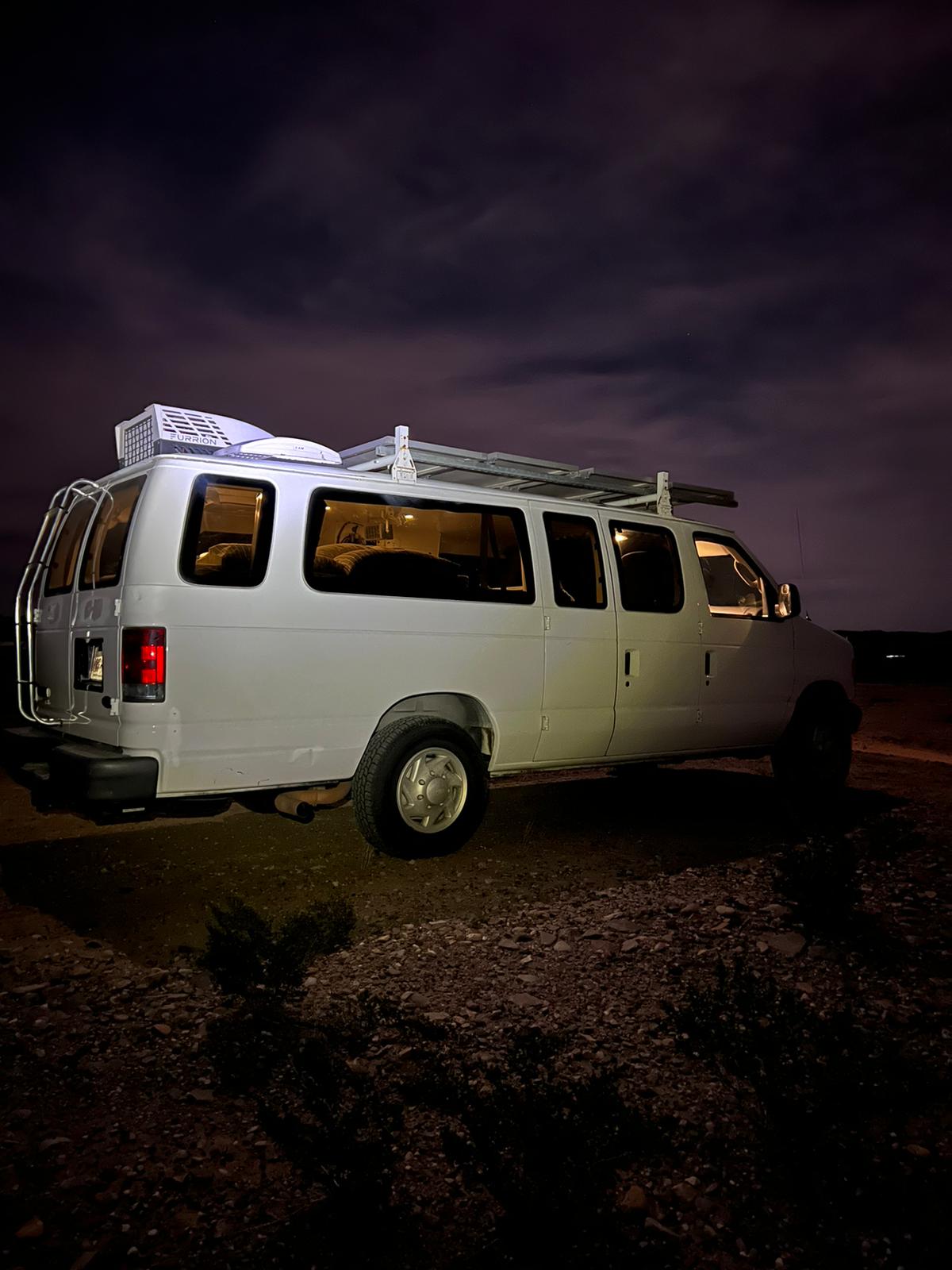 Night shot of a white camper van with a rooftop AC & roof rack. Ideal for an RV rental adventure under cloudy skies.