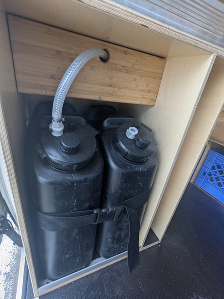 RV rental: Interior storage featuring two black water tanks connected by a hose, secured within custom wooden cabinetry.