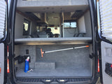 Spacious RV rental interior: two-tier setup with lower carpeted cargo area holding a broom and hose, and an upper bed/storage platform.
