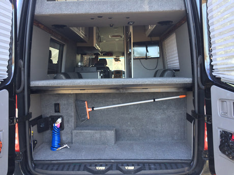 Spacious RV rental interior: two-tier setup with lower carpeted cargo area holding a broom and hose, and an upper bed/storage platform.