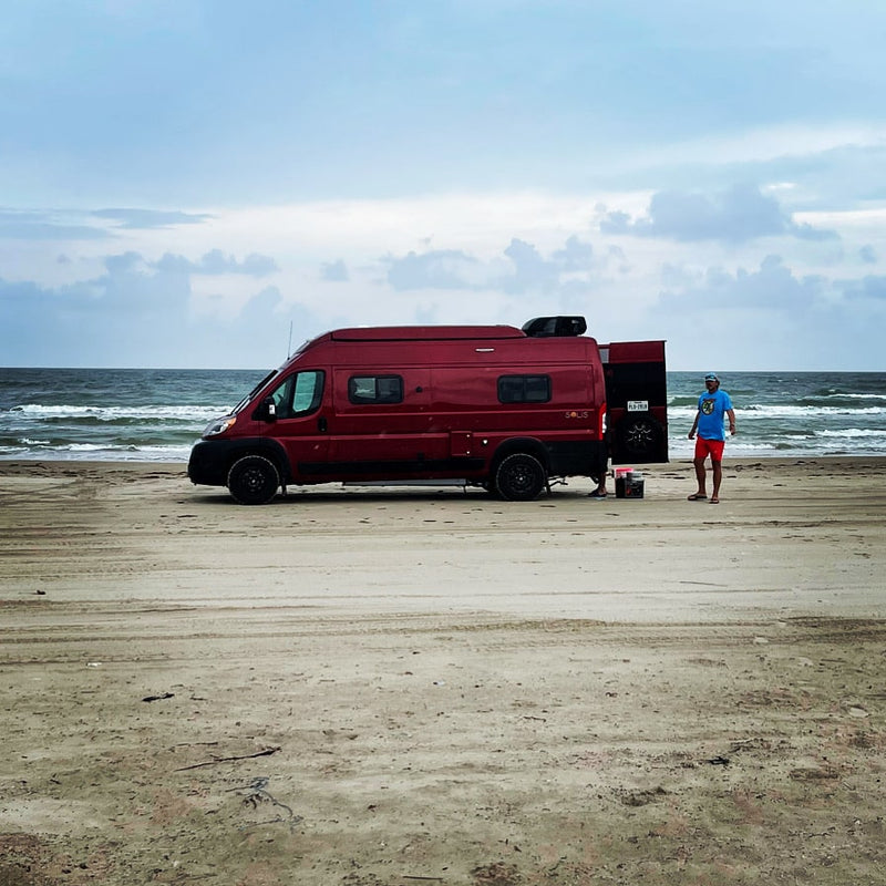 Red RV rental camper van parked on a sandy beach with ocean waves. A person is near the open rear door, enjoying beach camping. — photo 7