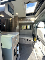 View of a modern RV rental interior showcasing kitchen, living area with bench seat, and bright overhead skylight.