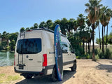 Experience an RV rental adventure! This light-colored camper van, with a paddleboard, is nestled among palm trees by water.