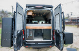 Custom RV rental van interior with open rear doors showing bed, utilities, and exterior storage boxes.
