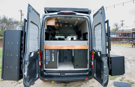 Custom RV rental van interior with open rear doors showing bed, utilities, and exterior storage boxes.