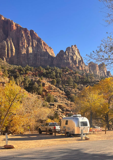 A shiny RV rental and truck parked at a vibrant fall campsite, beneath stunning red rock cliffs and a clear blue sky.