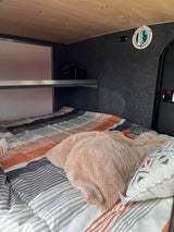 An inviting rv rental sleeping area, featuring a striped bedspread, soft blanket, wooden ceiling, and a practical shelf unit.