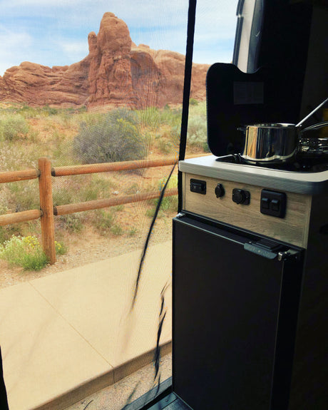 RV rental interior showing a compact camper kitchen, with a stunning desert view of red rock formations through a mesh screen.
