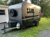 Dark gray "Flyer" teardrop RV rental, equipped with a spare tire & propane, parked on gravel for your next adventure.