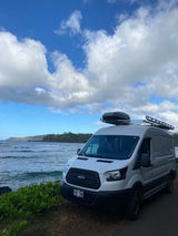 White Ford Transit rv rental parked on Kauai's scenic ocean shore, equipped with a roof box for adventure.