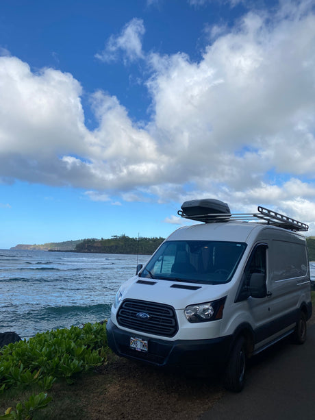 White Ford Transit rv rental parked on Kauai's scenic ocean shore, equipped with a roof box for adventure.