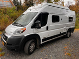 White Winnebago Solis camper van RV rental on gravel, surrounded by autumn foliage and a building.