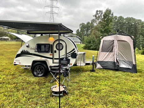 RV rental: A white teardrop camper with an awning and attached tent, campfire, and chairs on a grassy field, ready for camping.