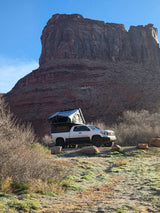 Overlanding RV rental: White pickup with rooftop tent & camper shell parked by towering red rock cliffs on frosty ground.