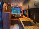 Step inside this charming RV rental: a rustic wooden camper van conversion featuring a bed, kitchen, and ambient lighting.