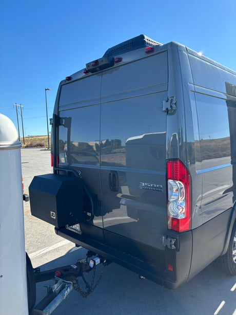 Rear view of a grey Ram ProMaster 3500 camper van, possibly an RV rental, with a tow hitch and storage box.