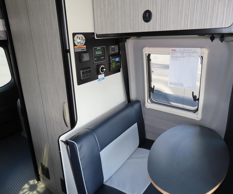 Inside an RV rental: a bench seat, round table, control panel with gauges & switches, plus a window & cabinet.