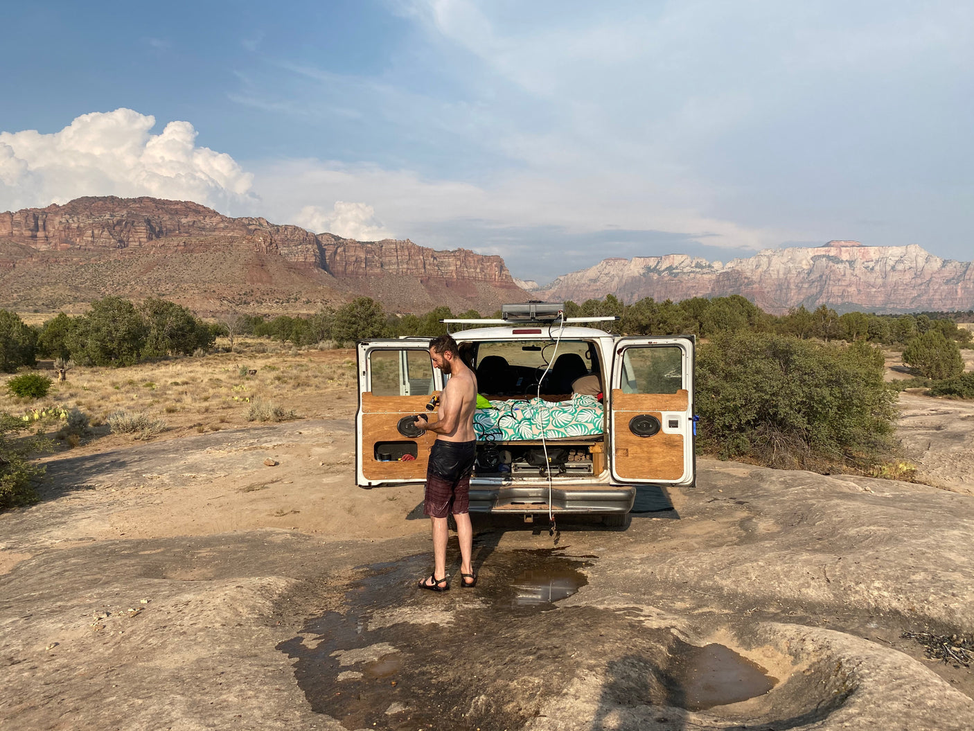 A man takes an outdoor shower next to his white rv rental van, set against stunning desert mountains. Ideal for camping.