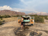 A man takes an outdoor shower next to his white rv rental van, set against stunning desert mountains. Ideal for camping.