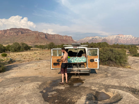 A man takes an outdoor shower next to his white rv rental van, set against stunning desert mountains. Ideal for camping.