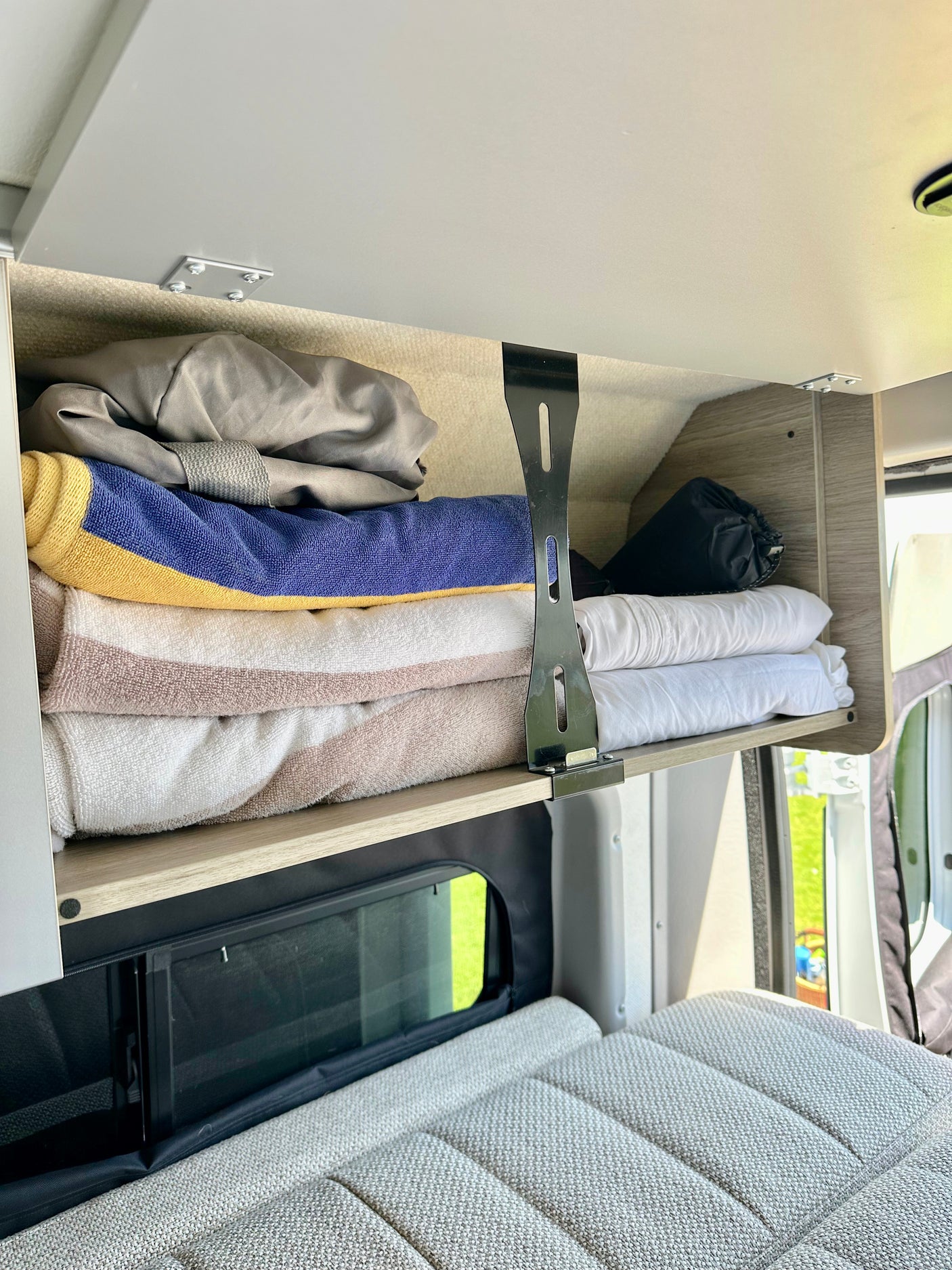 RV rental overhead cabinet with stacked towels and bedding, above a comfy grey bed and window.