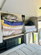 RV rental overhead cabinet with stacked towels and bedding, above a comfy grey bed and window.