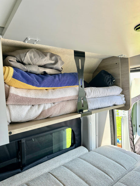 RV rental overhead cabinet with stacked towels and bedding, above a comfy grey bed and window.