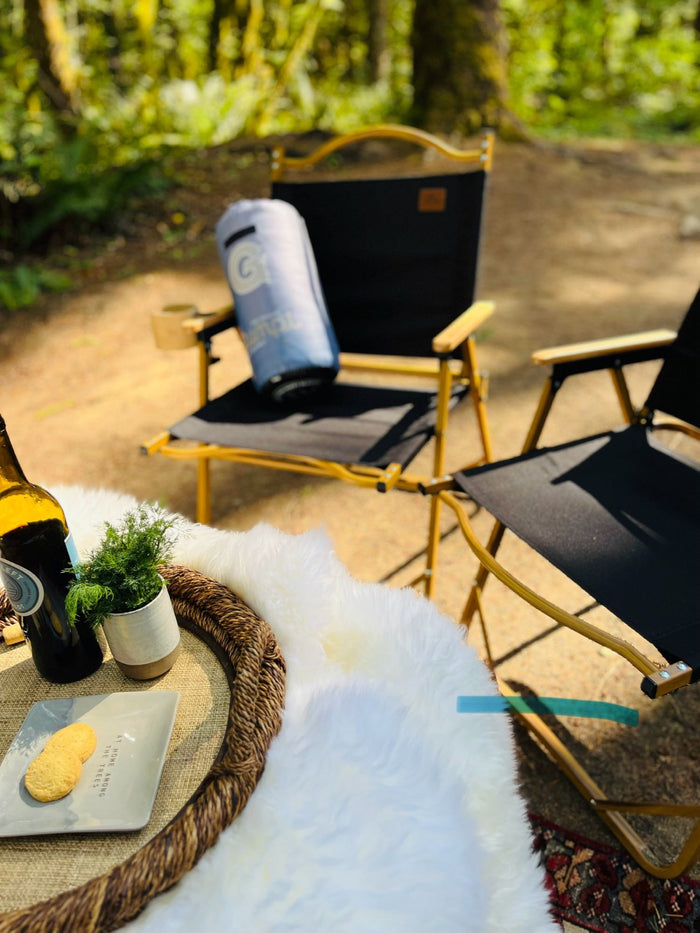 Campsite comfort with your RV rental! Two chairs, picnic setup, and a fluffy rug under trees for outdoor relaxation.