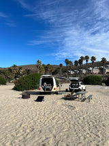 Beach camping with an RV rental setup. A person works on a laptop in the camper, surrounded by sand, trees, and blue skies.
