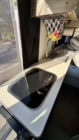 RV rental camper van interior showing a compact kitchenette with stovetop, sink, fruit basket, and outdoor window view.