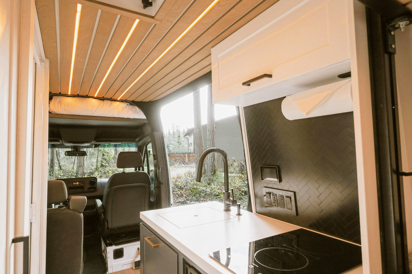 Stylish RV rental interior: modern kitchen, black herringbone backsplash, wood slat ceiling with LED lights, and bed.